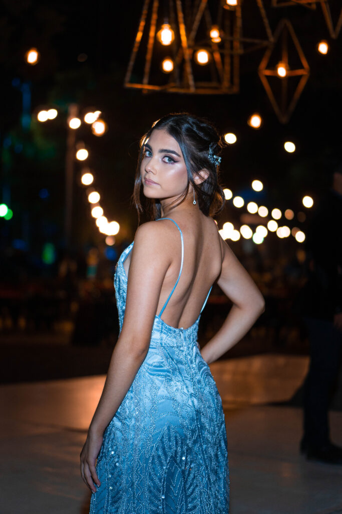 Egresada de espaldas con vestido celeste bajo guirnaldas de luces